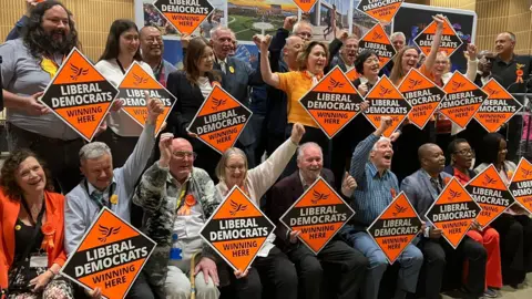 Two rows of Lib Dem councillors and supporters hold diamond shaped placards with orange and black colours that read: Liberal Democrats winning here. they are cheering and fist pumping. 