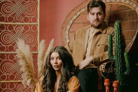 Smith & Liddle Billy Smith is sat on a wicker chair and Elizabeth Liddle is sat in front of him next to a cactus on a table.