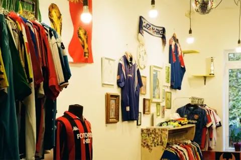 Polina and Serge The inside of the couple's vintage football shirt shop in Kyiv. On the left there is a rack of colourful football shirts, on the right is another two racks of colourful football shirts. In the centre of the image is the red and black striped shirt of AC Milan. There are shirts, scarves and flags and photos on the wall, with wire lightbulbs dropping from the ceiling.