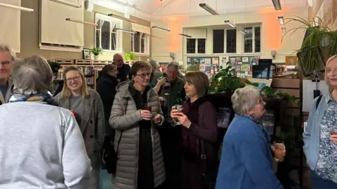 A group of people gathered in a library, chatting, at night. They are holding drinks and standing in little huddles of two and three. 