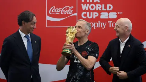AFP via Getty Images Canada's Prime Minister Mark Carney, in a black Canada jersey, kisses the FIFA World Cup trophy, as FIFA President Giovanni Infantino and Italian football coach Alessandro Nesta, both in suits, look on, amused. 
