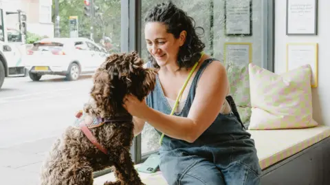 Vicki Shenkin Kerr and her dog sit in her bookshop window. Ms Shenkin Kerr  wears denim dungarees. She has dark, curly hair. Her dog has brown, curly fur.