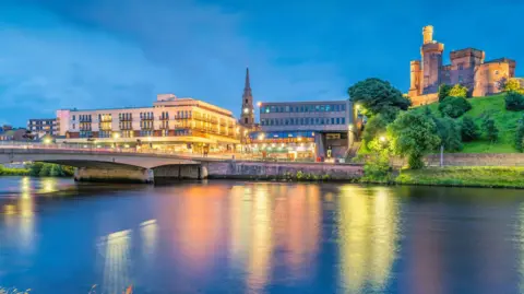 Buildings on Inverness' riverside are lit up at night. The buildings include a large block of flats, the city centre's steeple and Inverness Castle. The lights are reflected in the River Ness.