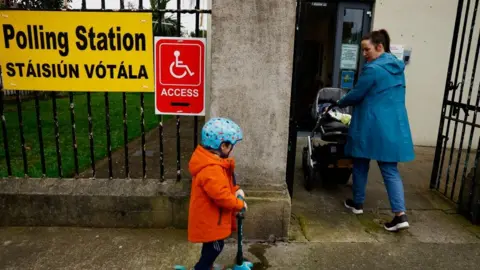 Ronald Grant Mother pushing pram with child on scooter behind her entering polling station