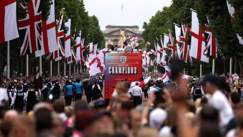 Getty Images An open-top England women's victory parade bus, displaying "EUROPEAN CHAMPIONS" on the back, driving down The Mall in London. The tree-lined street is heavily decorated with Union Jacks and St George's Cross flags, with a large crowd watching and Buckingham Palace visible in the background.
