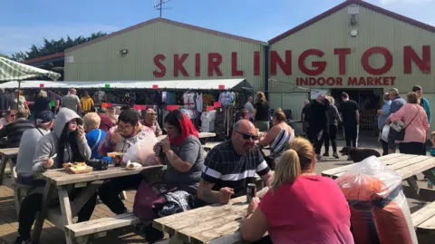 Skirlington Market Several benches located in an outside seating area with several people sat on them with canned drinks and food. In the background is a warehouse-type building which says 'Skirlington Indoor Market' in large red letters.