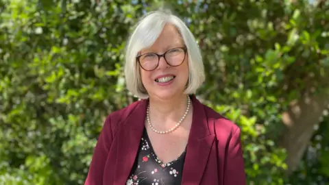 Dr Nicola Brink stands outdoors on a sunny day, wearing a burgundy jacket and glasses, with greenery in the background behind them.