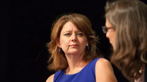 Mogan Selvakannu Close up of Leanne Wood looking like she is listening to another woman speaking during a debate. She is wearing blue dress and is against a black back drop