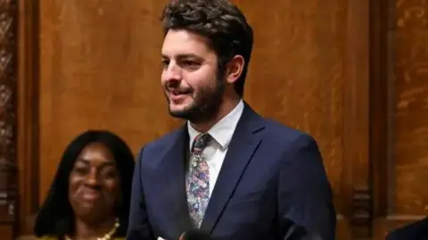 Ipswich MP Jack Abbott speaks in Parliament in front of wooden-panelled background, wearing a blue suit, white shirt and patterned tie.