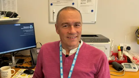 Josh McLaughlin/BBC Dr Andrews sits at his desk in his consulting room. He has short cropped dark hair. He wears a bright pink jumper and his blue NHS lanyard.
