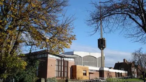 Oxymoron External view of the church, showing a tall 'spike' in front of a series of brick and concrete oblong buildings