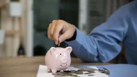Getty Images Man putting a coin into a pink piggy bank