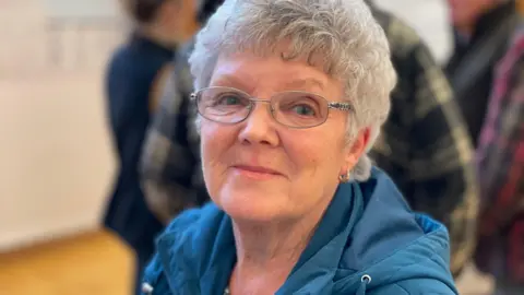 A woman with short grey hair wearing a blue coat and glasses. She is standing in a room full of people, who are standing in the background out of focus. 
