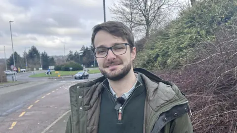 BBC/ Kate Bradbrook Man in his thirties with brown hair, beard and glasses standing by the side of a road. He is wearing an olive green coat and green sweater.