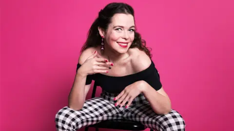 Juliette Burton Juliette Burton sitting on a black chair wearing black gingham trousers and a black off-the-shoulder top, posing in front of a shocking pink background