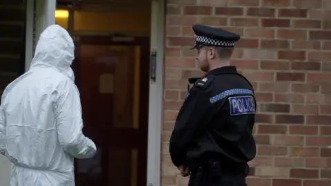 A police man wearing black uniform. He has his back to the camera and is facing the entrance of  the flat that has its front door open.
A second person is wearing a white forensic suit standing to the left of the policeman.