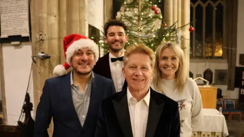 Four people stood next to each other. Three men are dressed in suits and the women is wearing a Christmas jumper