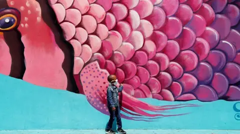 A boy wearing a protective face mask touches a wall of graffiti