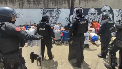 Ministry of the Interior of Ecuador Heavily armed police guarding inmates on the ground with their hands behind their heads.