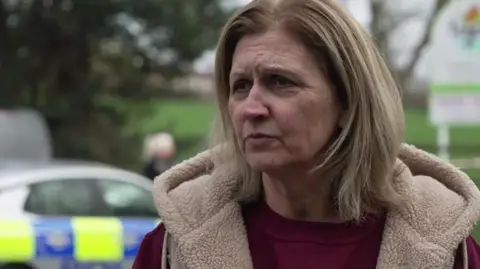 BBC Alison Huntley owner and founder of Tigers Day Nurseries - she has blond hair and wears a dark red top with a light brown jacket - a police car is behind her.