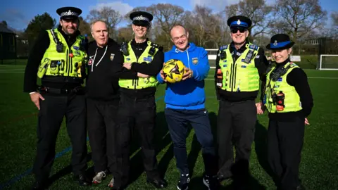 Getty Images Liberal Democrat leader Ed Davey stands alongside a group of police officers in uniform. He is holding out a yellow football and they are all stood on a football pitch at Wiltshire FA headquarters in Devizes