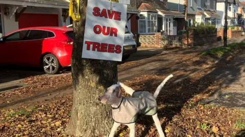 A tree with a sign and a dog ornament in front. The sign on the tree reads 'Save our trees' in red lettering.