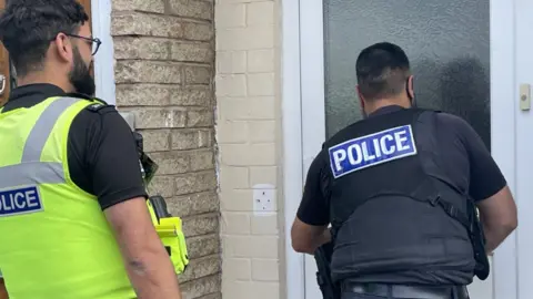 Two uniformed police officers approach a white front door. One officer is in a fluorescent jacket, the other black. Both have their backs to the camera.