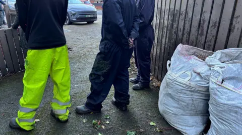 Three men stand with their back to the camera. The two men to the right wear identical navy boiler suits and black boots. The man to the left wears hi-viz trousers and a navy jumper, with black boots. There are tow large white bags behind them resting on a brown fence. 
