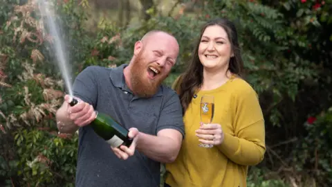 National Lottery Craig opening a bottle of champagne and spraying it everywhere whilst smiling, and Zoe holding a glass of champagne and also smiling.