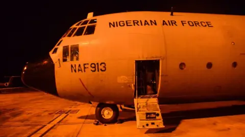 Getty Images The nose of a Nigerian air force plan on the runway. The photograph is taken at night and the plane is bathed in an orange light.