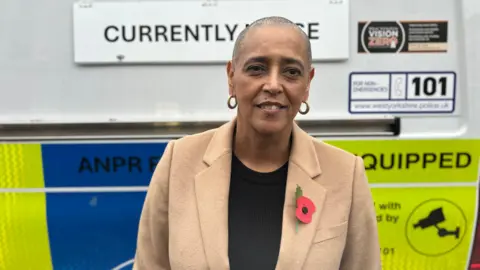 Beth Parsons/BBC Lowe stands in front of a police vehicle that displays signage related to surveillance technology. The van behind her has a large sign reading “FACIAL RECOGNITION CURRENTLY IN USE”, along with other markings indicating police contact information and safety initiatives. The vehicle also features blue and yellow police livery with the phrase “ANPR EQUIPPED", referring to automatic number plate recognition. Lowe is wearing a beige coat over a black top and has a red remembrance poppy pinned to her lapel. Gold hoop earrings are visible.