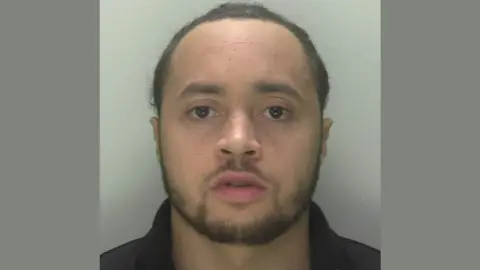 A police custody photo of Kiano Houghton Jones. He has short dark brown hair, a dark brown beard and is wearing a black collared top.