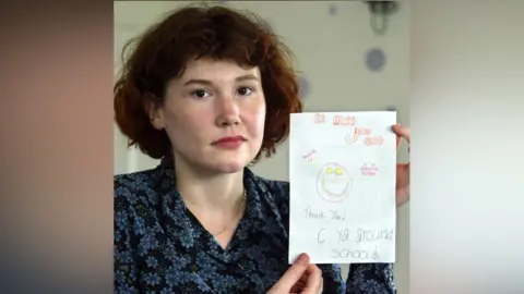 PA Media Maxine Carr: A woman with pale and red hair holding a handwritten card next to the left side of her head. She is wearing a blue shirt with a floral pattern.