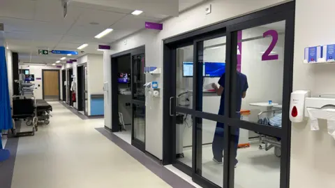 Corridor of a ward with glass sliding doors to cubicles. The back of a member of staff can be seen at a computer. 