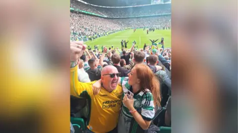 A man wearing a yellow Celtic jersey is smiling and raising his fist in the air. He has his arm around a young woman who is also smiling and wearing a green and white Celtic jersey. They are standing in a football stadium and behind them lot of fans are celebrating. 