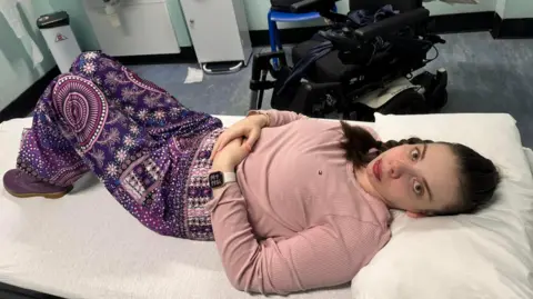 Katie Robertson A woman wearing a pink jumper and purple patterned trousers lies on a hospital bed with a wheelchair in the background