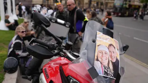 PA Media. Una imagen de Craig y Lindsey Foreman sonriendo, pegada con cinta adhesiva en la parte delantera de una motocicleta roja de gran tamaño. Varias personas y otras motocicletas se pueden ver al fondo, en lo que parece ser la Plaza del Parlamento en Londres.