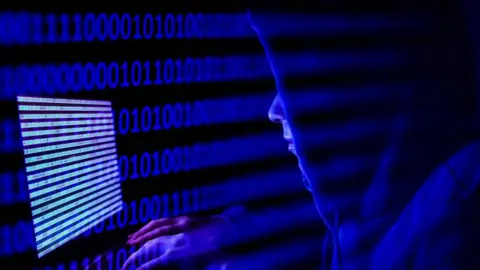 Getty Images A person dressed as an internet hacker is seen with binary code displayed on a laptop screen.