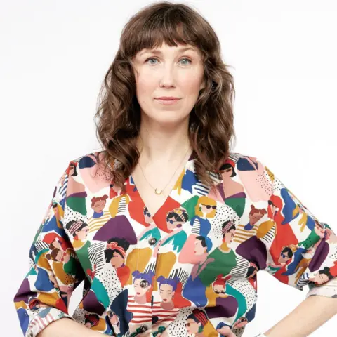 Meagan Shuptar Bergen Anderson looks into the camera posing for a photo. She has shoulder-length brown hair and a fringe. She is wearing a blouse featuring illustrations of women on it.