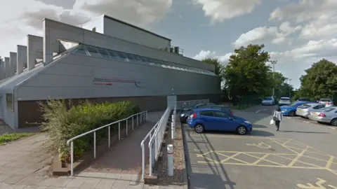 Google The outside of Bury St Edmunds leisure centre. A large white building with red and black writing on, Welcome to Bury St Edmunds Leisure Centre, with a sign which says reception and an arrow pointing to the right side of the building. There is also a car park outside the leisure centre with disabled parking bays and a ramp up to the leisure centre with white railings.