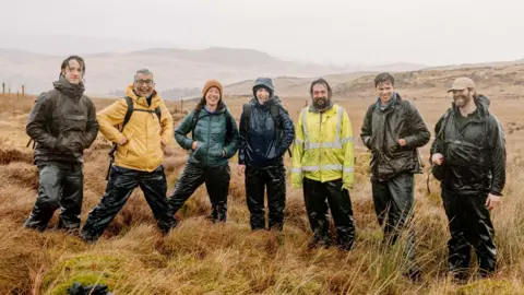 Five men and two woman wearing waterproof outerwear, smiling and standing on open grassland.