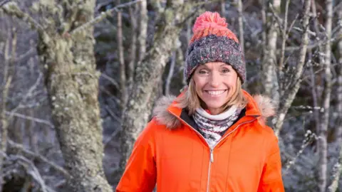 A smiling Michaela Strachan posing for the camera, wearing a padded orange jacked and a multicoloured woolly bobble hat.