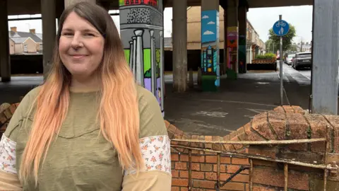 A woman in a green top stands in front of a wall with large pillars visible behind her.