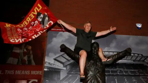 Reuters A Liverpool fan celebrates winning the Premier League as he sits on a statue of Bill Shankly outside Anfield