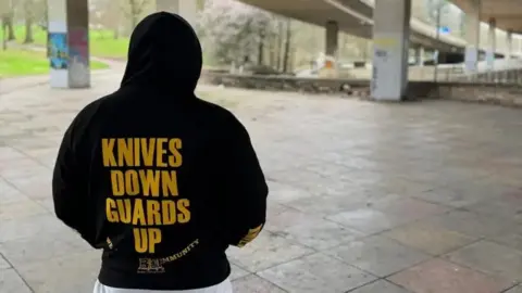 A young person faces away from the camera. They are wearing a black hoodie with "Knives Down Guards Up" in bronze capital letters on the back. They are standing in a paved area underneath a motorway