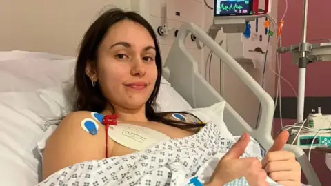 Faith Harrison A woman with long dark hair in a hospital gown lying in a hospital bed with both thumbs up