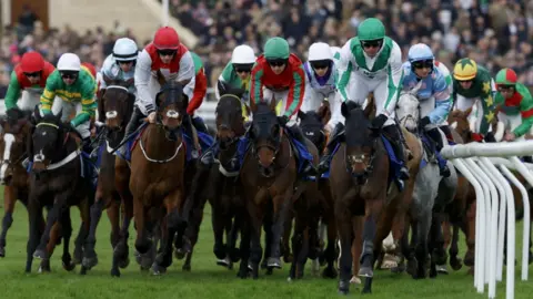 Peter Cziborra/Reuters A group of horses ridden by jockeys in multi-coloured silks turn a bend on the course. Behind them are grandstands packed with people.