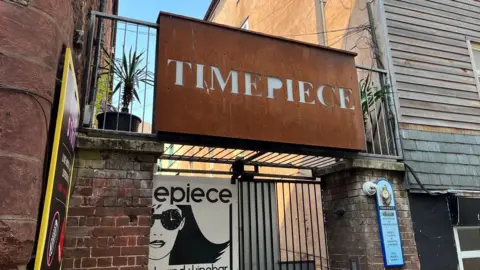 BBC An oxidised metal sign with Timepiece in white above the entrance to the Timepiece nightclub in Exeter. Other signs are on the surrounding walls and gates at the building.