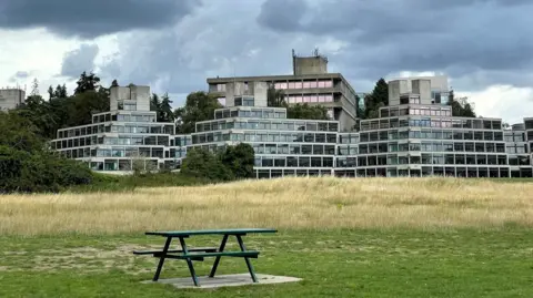 Shaun Whitmore/BBC The Ziggurats accommodation buildings at the UEA. They are seven storey concrete and glass buildings that rise like steps. A picnic bench on a patch of grass is in the foreground and the sky is overcast