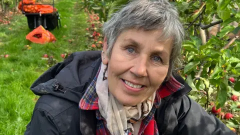 Roz Scott is pictured smiling at the camera. She is sitting in an apple field and has short grey hair with several ear piercings. She is wearing a black coat with a cream coloured scarf around her neck. To her right lies a line of apple trees. A basket filled with apples can be seen behind her. 